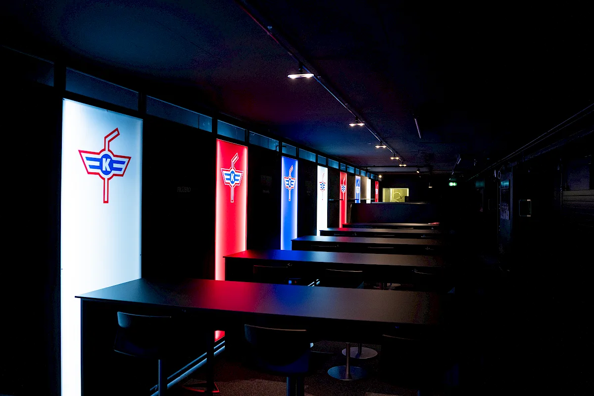 Dunkler Gang der VIP-Loge mit beleuchteten Vereinslogos und Stehtischen mit Barhockern