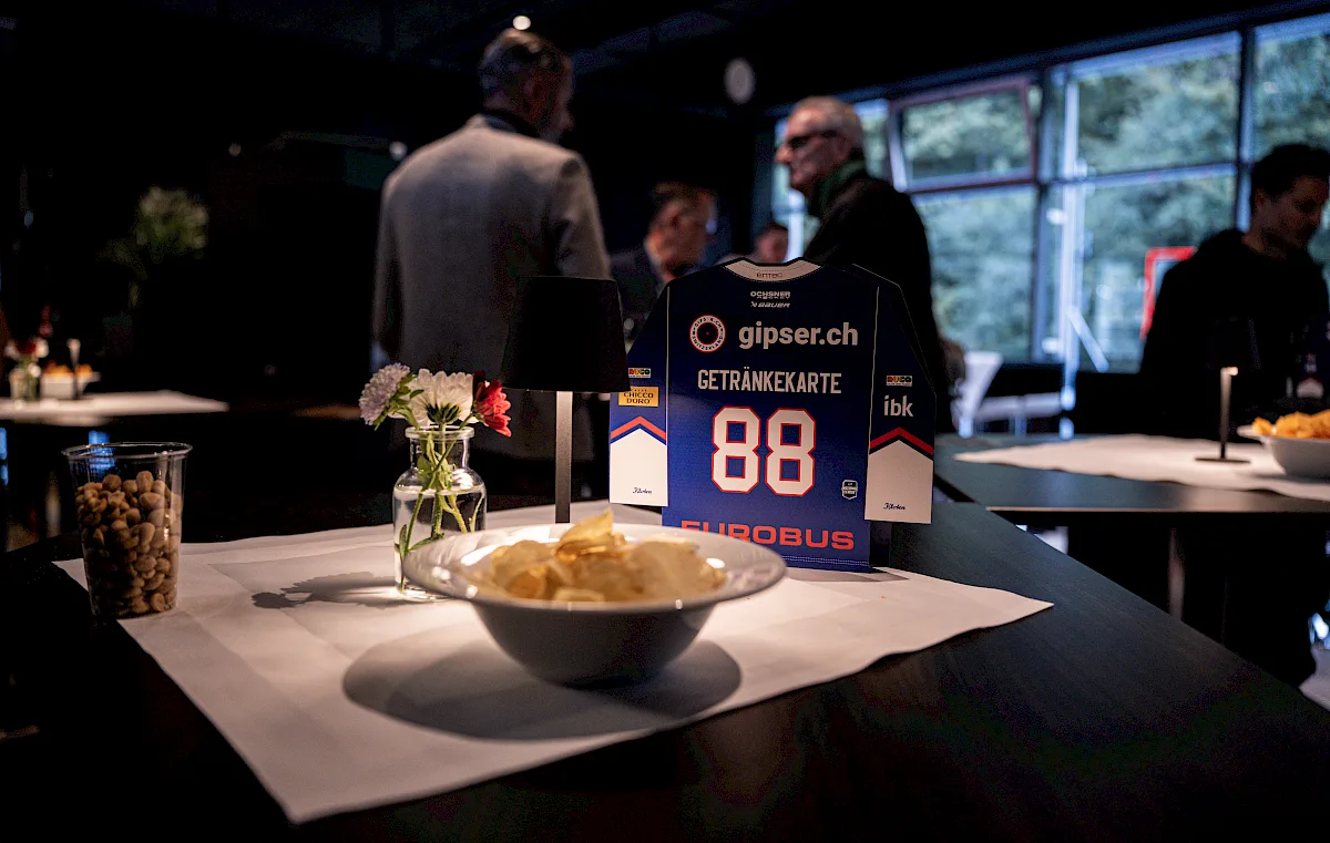 Getr&auml;nkekarte im Trikot-Design mit Snacks und Blumen auf einem Tisch, im Hintergrund G&auml;ste