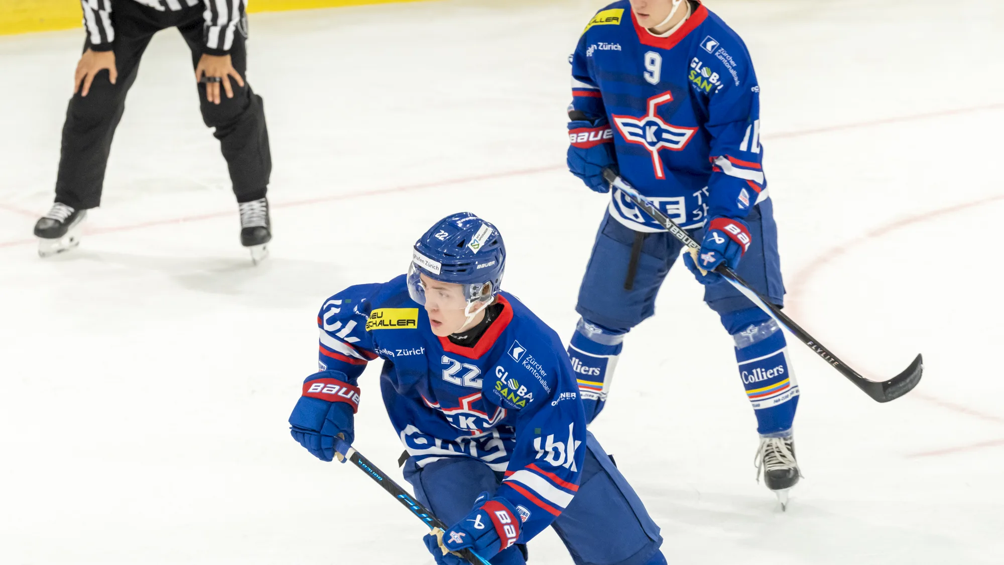 Zwei Spieler der U21-Elit in blauem Dress mit Puck auf dem Eis, im Hintergrund ein Schiedsrichter